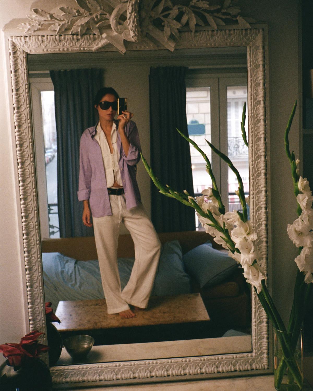 Diana Bartlett taking a self-portrait with a vintage-style camera, wearing dark sunglasses and a striped shirt in a softly lit interior with warm tones and elegant details.