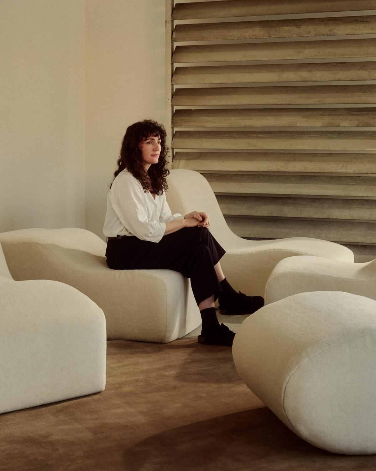 Porträtt av Sophie Pearce, sittandes i en skulptural krämfärgad fåtölj ovanpå mattan Grand i färgen Chestnut.