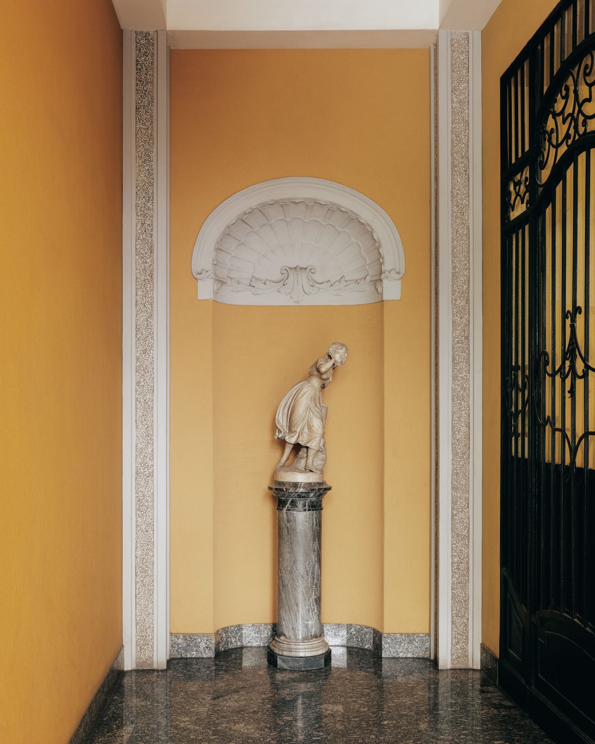 Ein historisches Interieur in Mailand mit einer klassischen Statue in einer gewölbten Nische, warmen Ockertönen und dunklem Marmorboden.