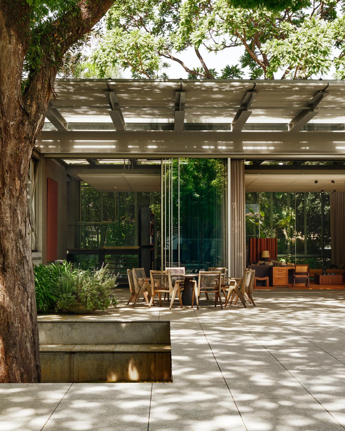 En modernistisk bostad i São Paulo, fotograferad från trädgården, där filtrerat dagsljus rör sig över en skuggad terrass med matgrupp i trä, inramad av grönska.
