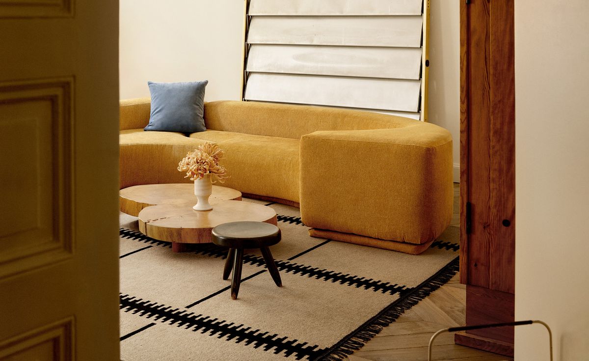 The Elder 06 rug in Black is shown in a contemporary living room with a mustard sofa, wooden coffee tables, and a blue cushion, creating a warm and modern atmosphere.