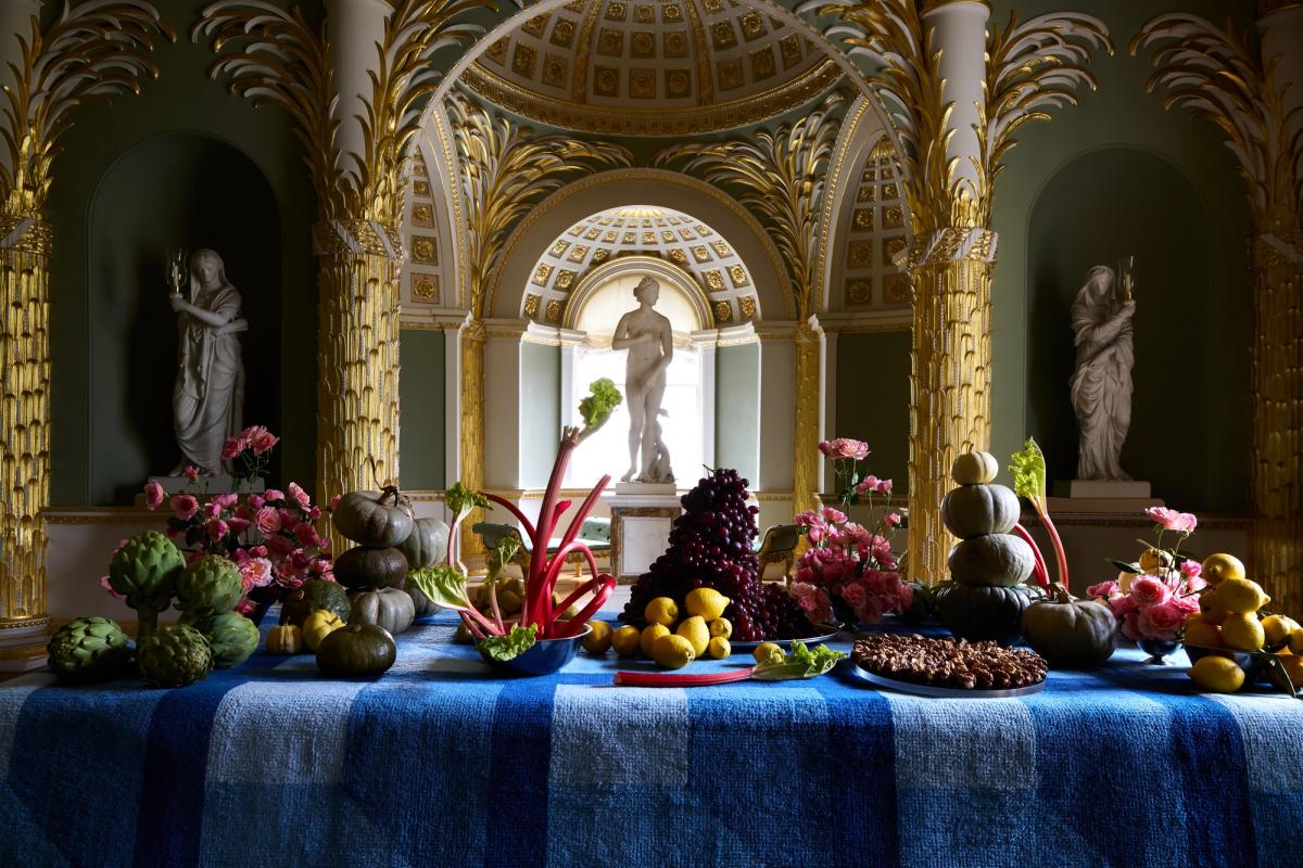 Plush wool rug Garden Maze in Blue pictured draped over a table with a decadent spread of fruits and flowers in a museum like setting.