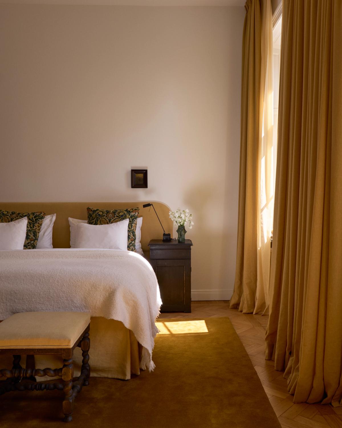 Ein warmes, elegantes Schlafzimmer mit dem Signature-Kopfteil in Leo, den Verdunkelungsvorhängen The Blackout in Leo und dem Teppich Grand in Leo, in goldenen Farbtönen.