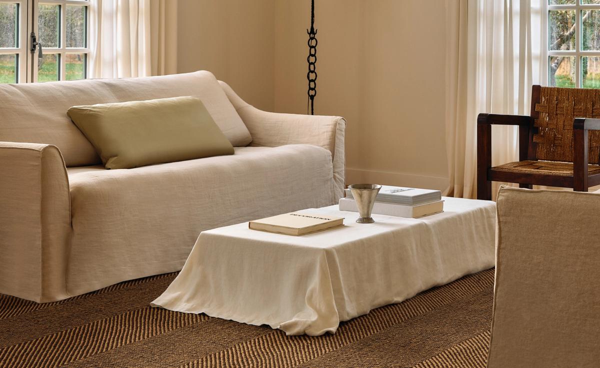 Jute Herringbone – Natural/Brown ancre un espace décontracté avec sa texture terreuse et son motif subtil. Un canapé recouvert de lin, un fauteuil d’appoint en bois tressé et une table basse drapée de tissu naturel reflètent la palette organique et apaisante de la pièce.