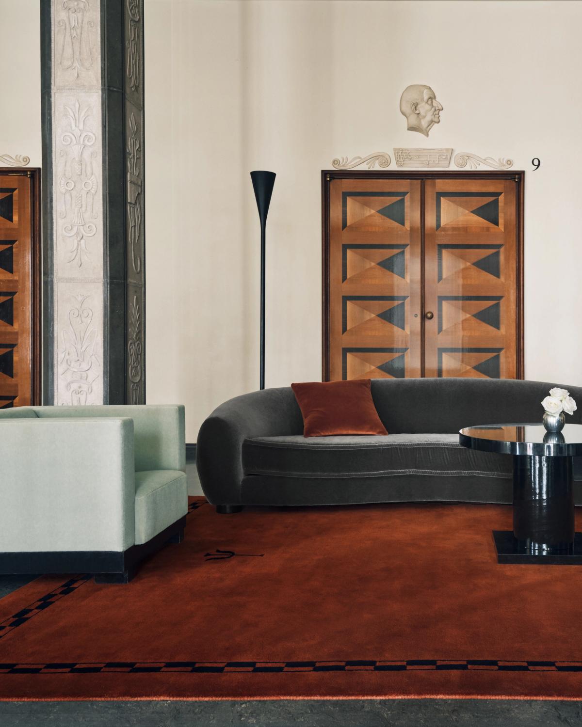 Grace 01 – Burgundy — styled inside Stockholm Concert Hall. A deep burgundy rug with a geometric border, paired with a grey curved sofa and mint green armchair.