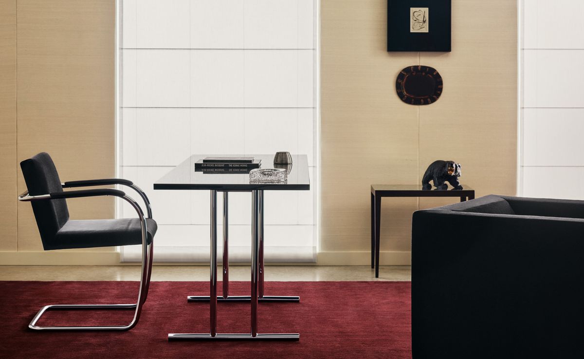 Der Teppich The Grand in Deep Wine, inszeniert in einem eleganten Interieur mit verchromten Stühlen, einem minimalistischen Tisch und warmen beigen Wänden für einen satten, ausdrucksstarken Kontrast.