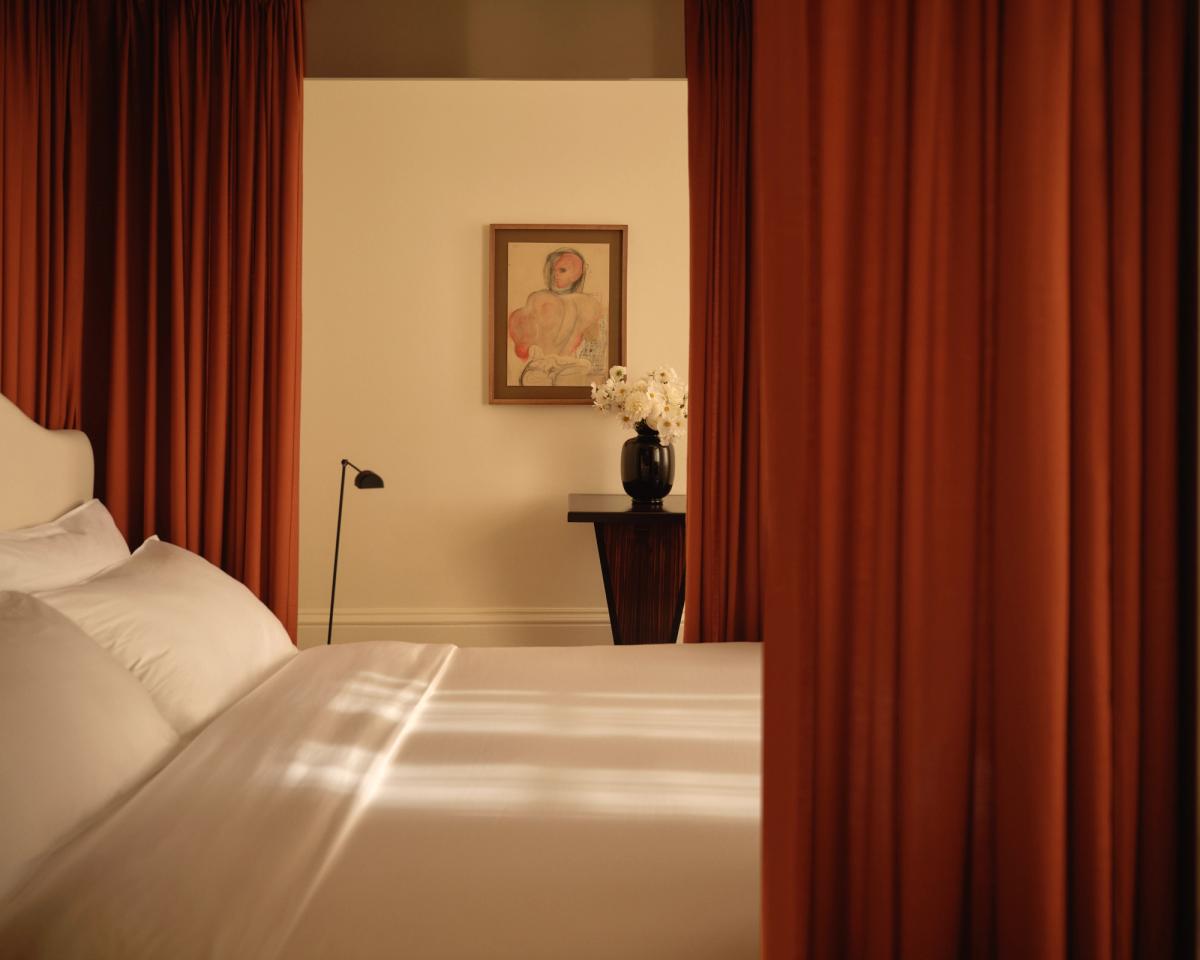 Ein ruhiges Schlafzimmer im Nordic Knots Store in der Mount Street in London. Frisch weiße Bettwäsche wird von The Grand Vorhängen in Burnt Red eingerahmt, während ein Beistelltisch aus dunklem Holz, ein Blumenarrangement und ein Kunstwerk dem Raum Wärme und Ausgewogenheit verleihen.