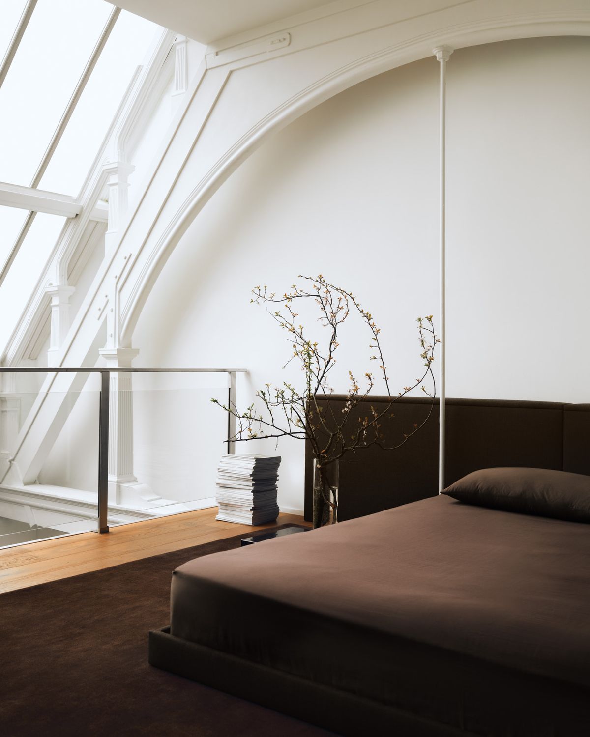 Der Teppich The Grand in Walnut, inszeniert in einem Loft-Schlafzimmer in Mailand mit einem niedrigen dunklen Bett unter einer weißen Gewölbedecke, kombiniert mit skulpturalen Sitzmöbeln und minimalem Dekor für eine warme, architektonische Atmosphäre.