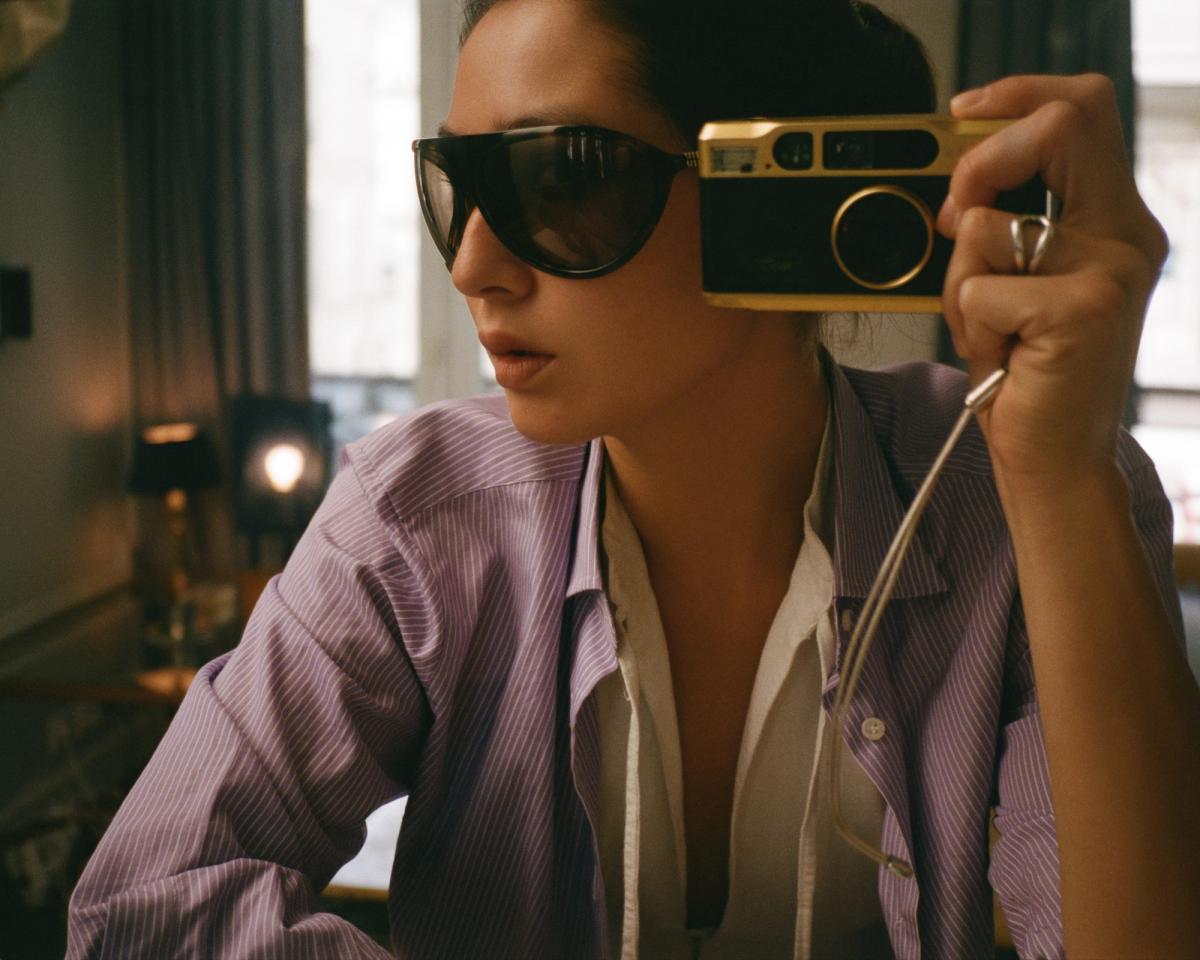Diana Bartlett macht ein Selbstporträt mit einer Vintage-Kamera, trägt dunkle Sonnenbrillen und ein gestreiftes Hemd, in einem sanft beleuchteten Raum mit warmen Tönen und eleganten Details.