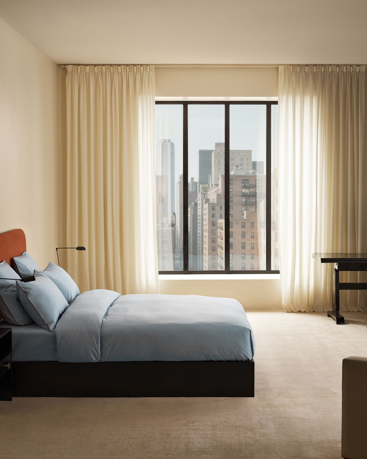 Ein modernes Schlafzimmer mit dem Signature-Kopfteil in Burnt Red, dem Percale-Bettwäscheset in Shirt Blue und den The Grand-Vorhängen in Pale Sand, mit Blick auf die Stadtsilhouette.