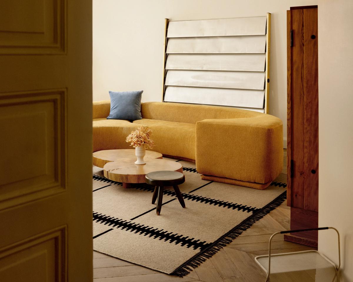 Salon chaleureux avec un canapé courbé couleur moutarde, des tables basses rondes en bois et le tapis Elder 06 – Black, orné d’une bordure linéaire audacieuse pour une touche graphique moderne.