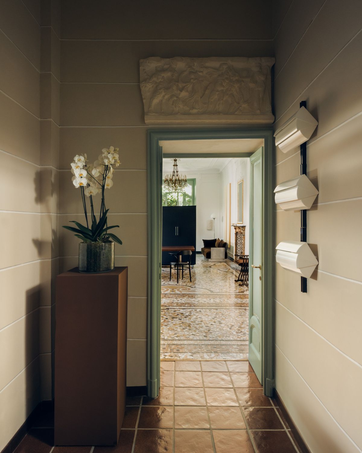 Ein schmaler Flur mit Fliesenboden und einem salbeigrünen Türrahmen, der in einen hellen Raum mit Terrazzoboden, Kronleuchter und skulpturalen Möbeln führt.