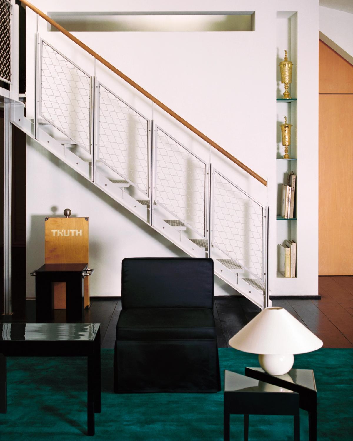 Grand – Emerald Green rug grounding a modern interior with a teal-green surface, black lounge chair, sculptural lamp, and a metal staircase in the background.