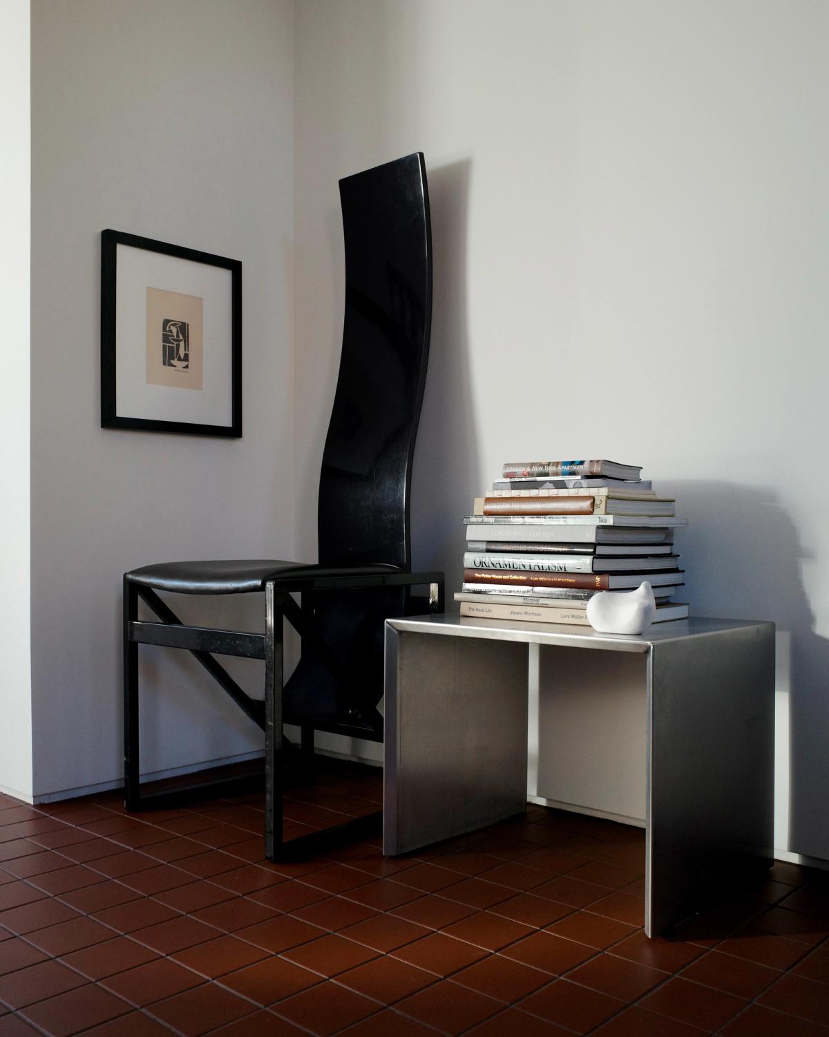 Photograph of a tall black artistic chair next to a metal table with a stack of books in a room with red tile floor at Monument Store.