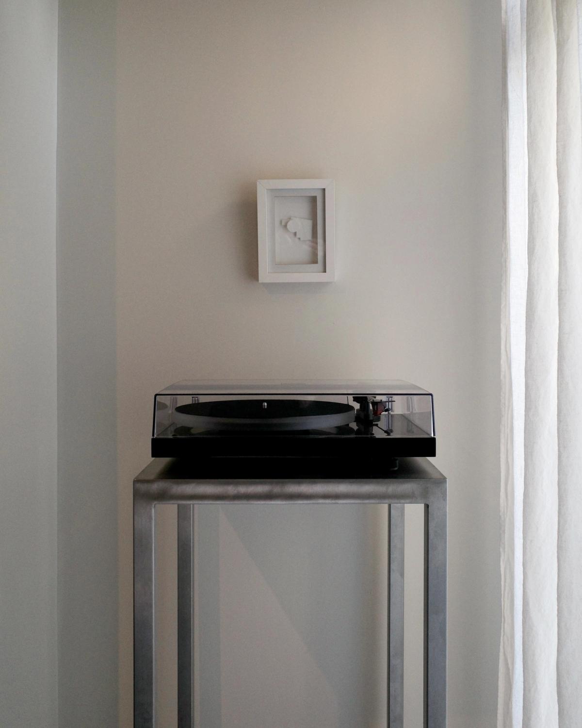 A photograph of a vinyl player on top of a small metal table next to a wall with an all white painting at Monument Store.