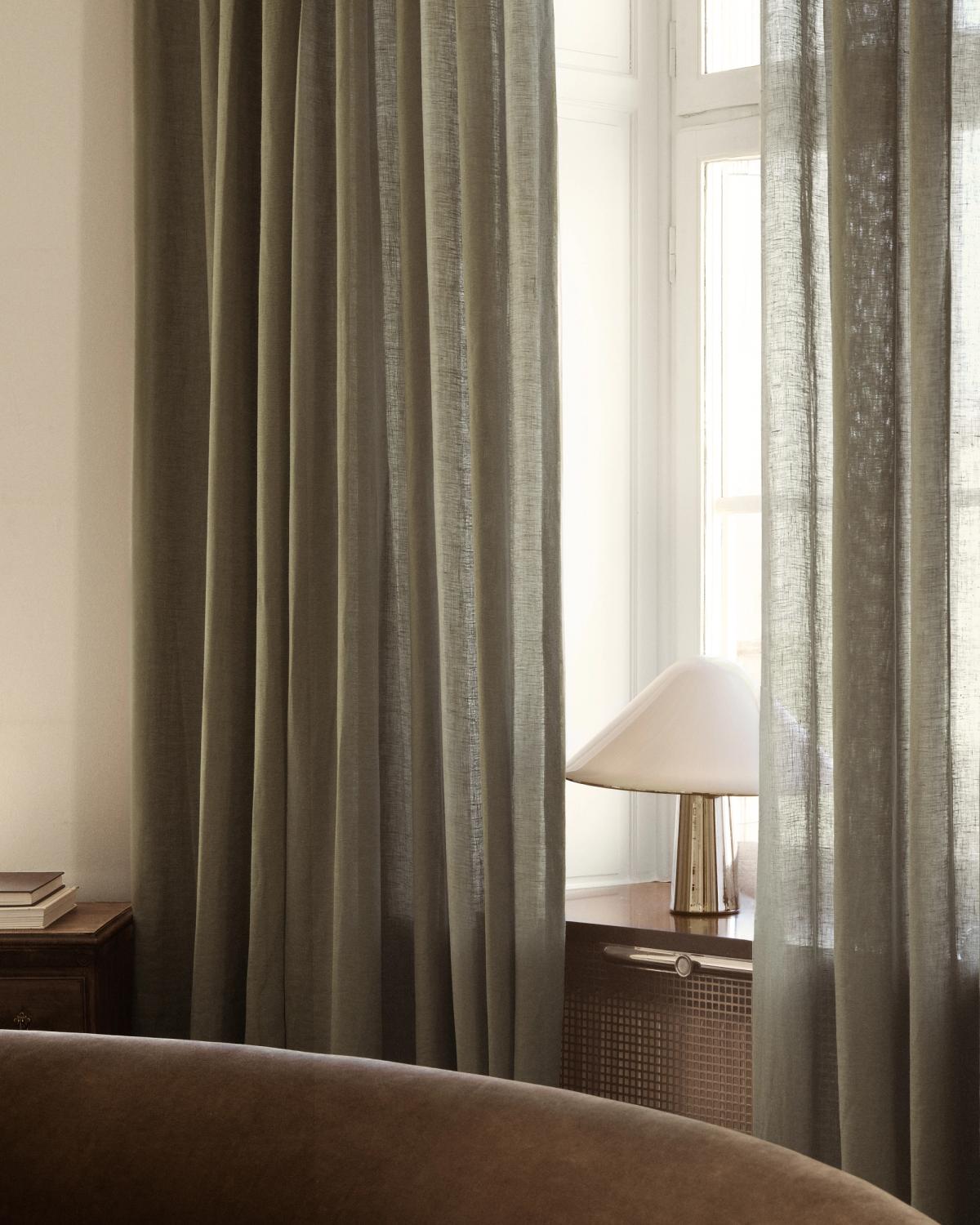 The Sheer Linen curtains in Olive in a softly lit room, framing a window with a sculptural table lamp and warm natural textures.