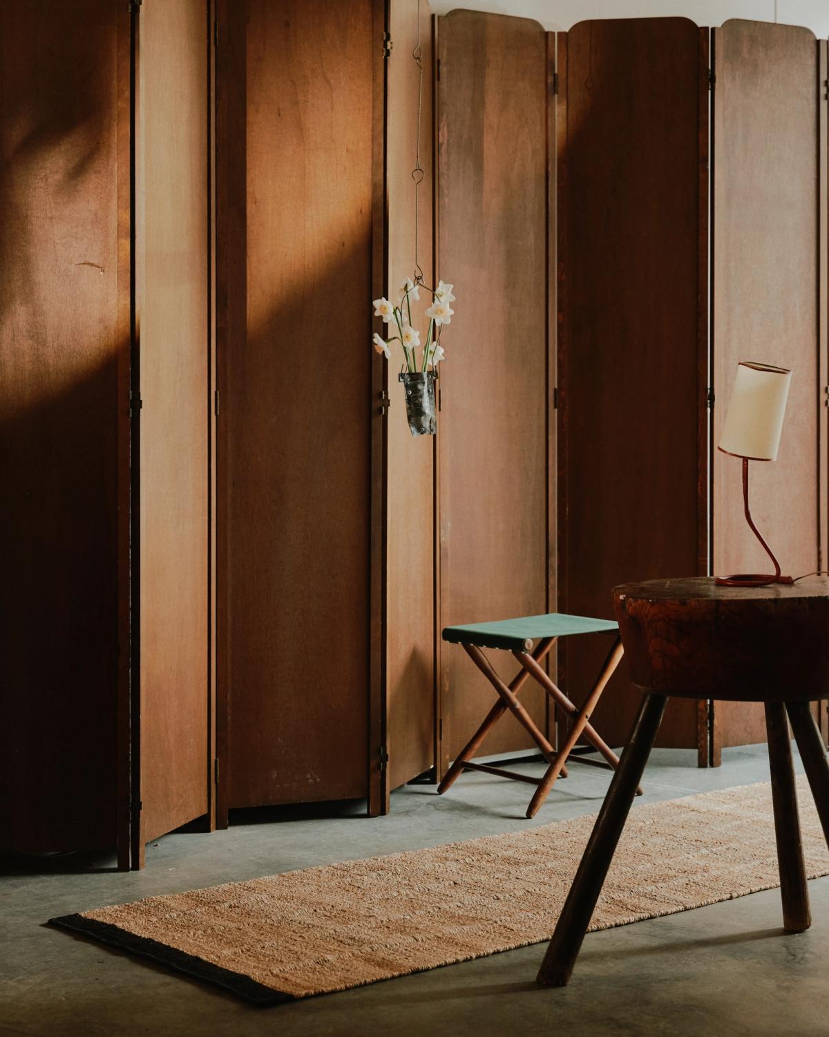 Jute Runner – Black adds a grounding, tactile presence to this warm-toned space. Its natural texture contrasts beautifully with the dark concrete floor and rich wooden folding screens, while sculptural accents and mid-century-inspired furniture complete the look.