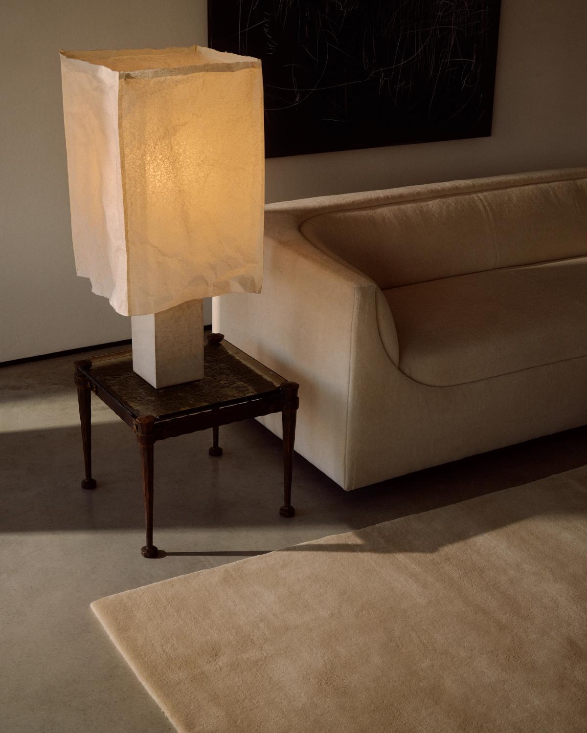 Närbild av mattan Grand i färgen Butter, intill en krämfärgad soffa och en skulptural lampa.
