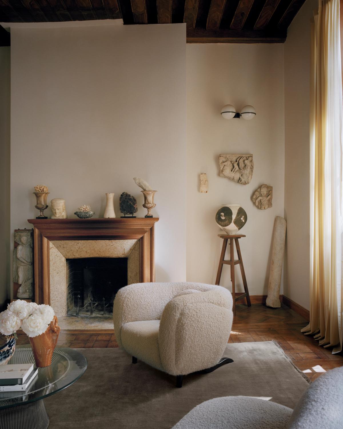 Le tapis Mohair en Taupe est présenté dans un salon parisien avec des fauteuils sculpturaux, une cheminée en pierre et une douce lumière naturelle, créant une atmosphère sereine et intemporelle.