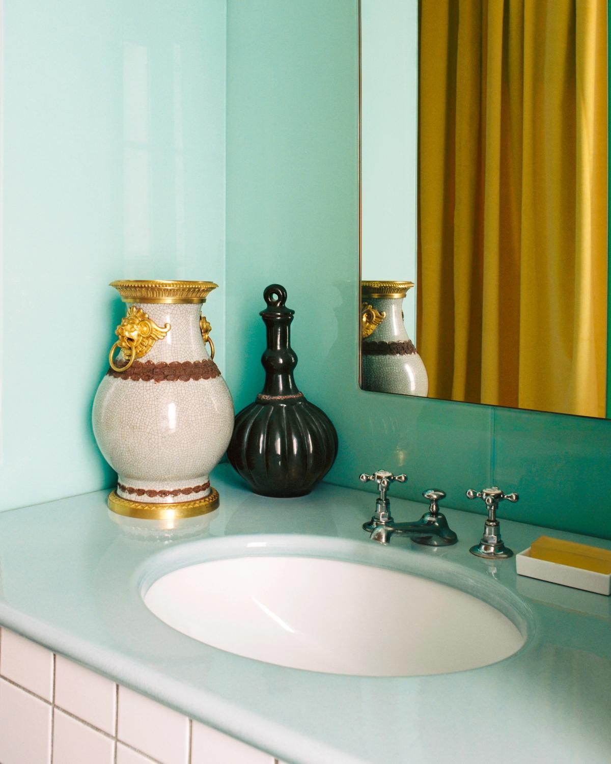 Bathroom vanity with a pale blue sink and mirror, styled with decorative vases, with The Grand curtains in Leo reflected in the background.