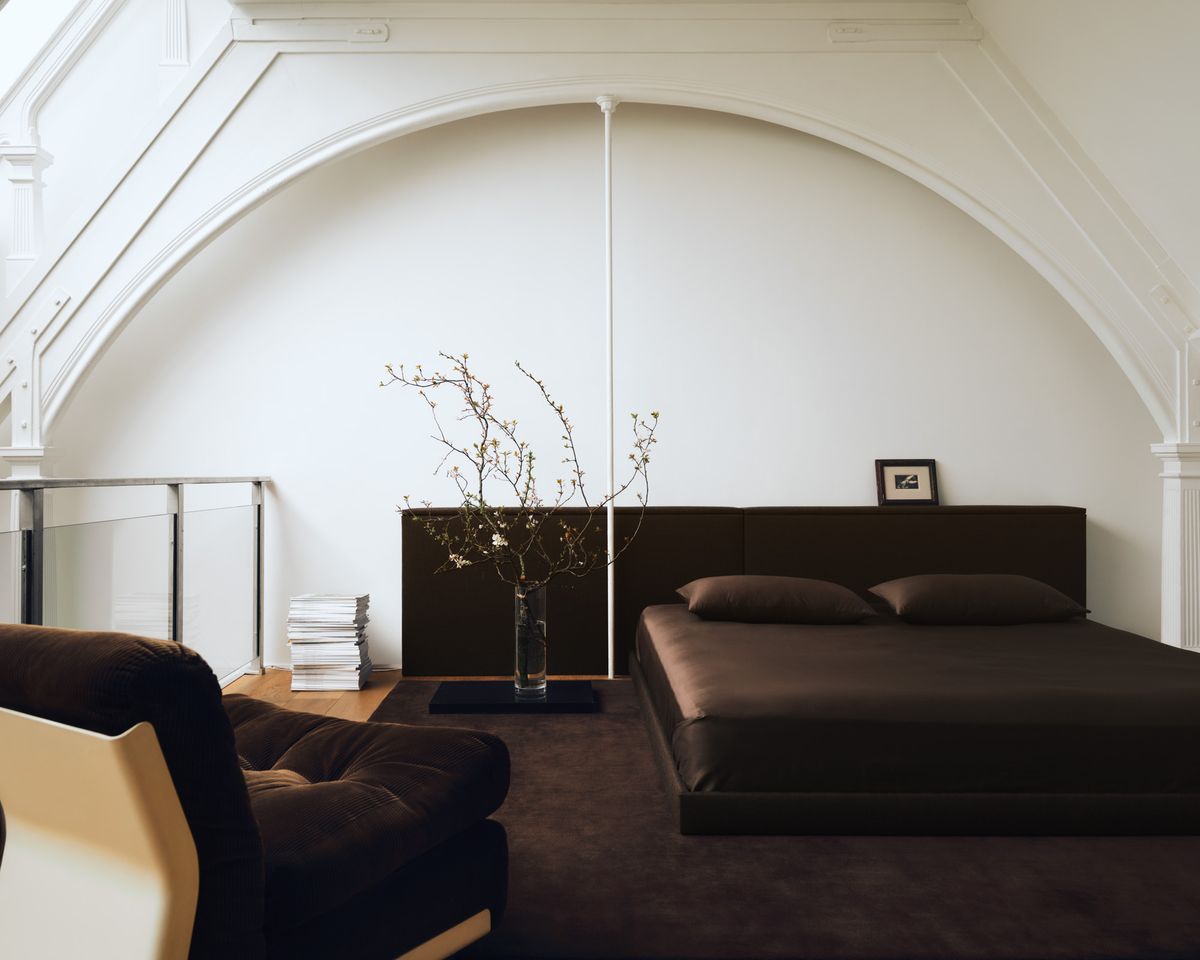 Der Teppich The Grand in Walnut, inszeniert in einem Loft-Schlafzimmer in Mailand mit einem niedrigen dunklen Bett unter einer weißen Gewölbedecke, kombiniert mit skulpturalen Sitzmöbeln und minimalem Dekor für eine warme, architektonische Atmosphäre.