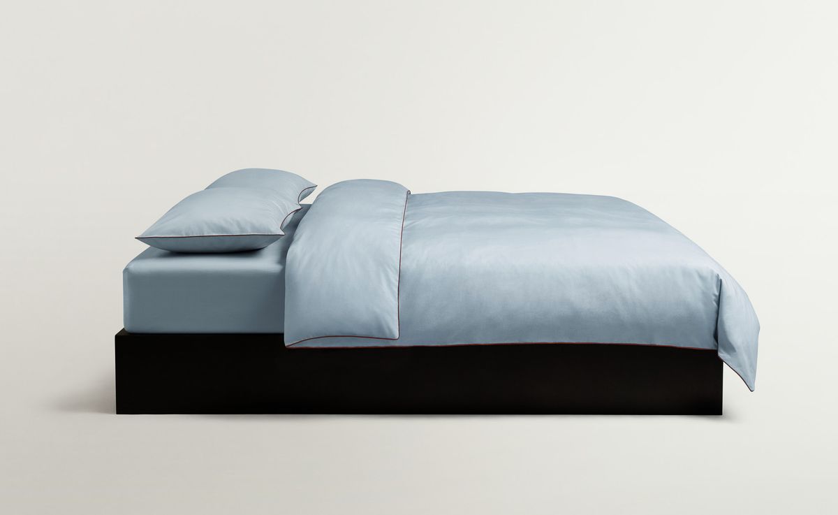 Percale Bettlaken-Set in Shirt Blue/Red, auf einem Bett arrangiert, aus hellblauer Baumwollperkal mit feiner roter Paspelierung für eine klare, maßgeschneiderte Optik.