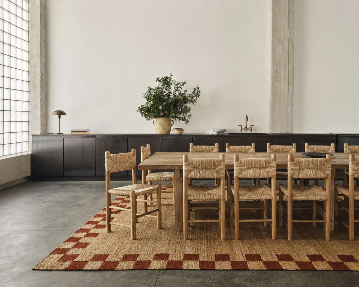 Un grand tapis Jute Border en Red, présenté sous une immense table à manger entourée de plusieurs chaises en bois et en rotin. L’espace est vaste et lumineux, avec une cuisine sombre à l’arrière-plan.
