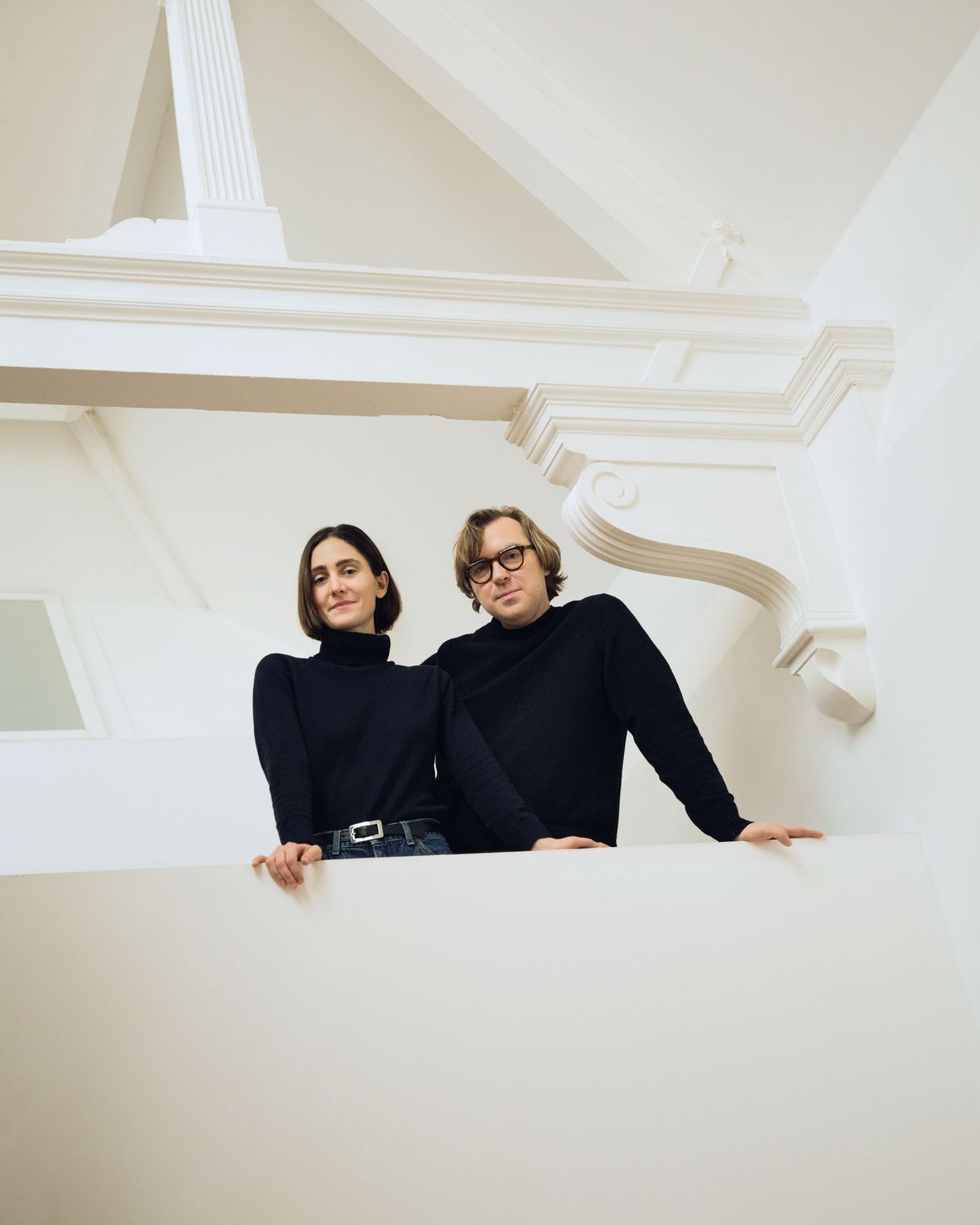 Porträt von Felizia Berchtold und Oskar Kohnen von Tutto Bene auf einem weißen Balkon, in Schwarz gekleidet, eingerahmt von ornamentalen architektonischen Details.