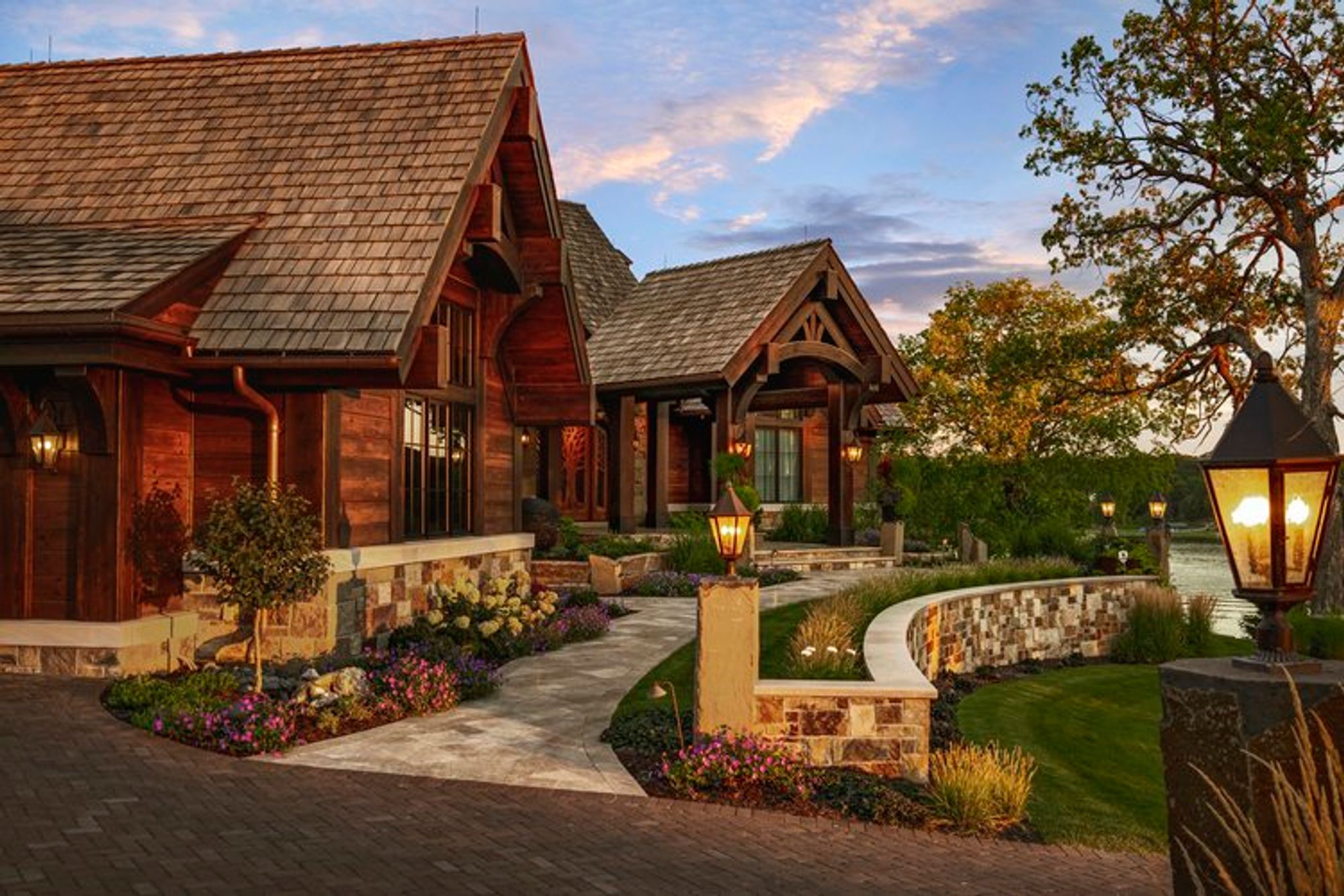 Lantern-lit stone piers and curved stone retaining wall beside a paver walkway, with a timber home and lake at dusk - Mom's Design Build