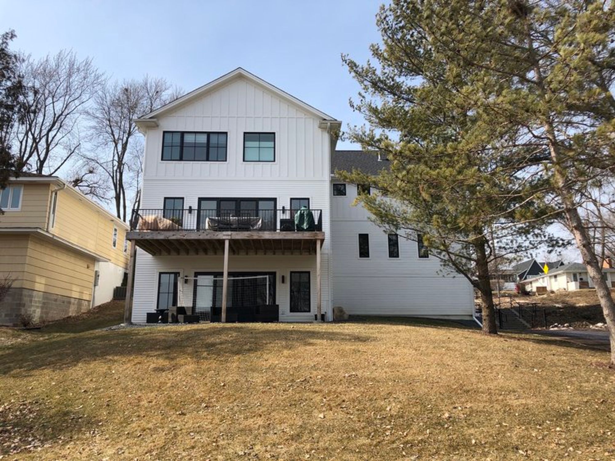 Rear of white board-and-batten home with upper deck and walkout lower level over a grassy slope - Mom's Design Build