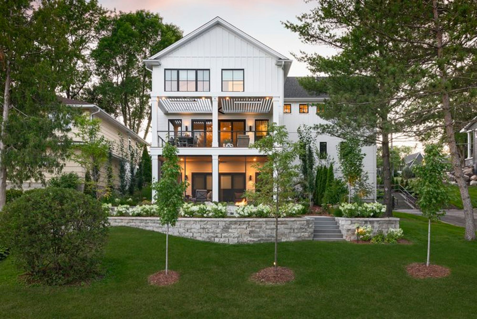 Rear of white home at dusk with lit porches, stone retaining wall, steps and hydrangea plantings - Mom's Design Build