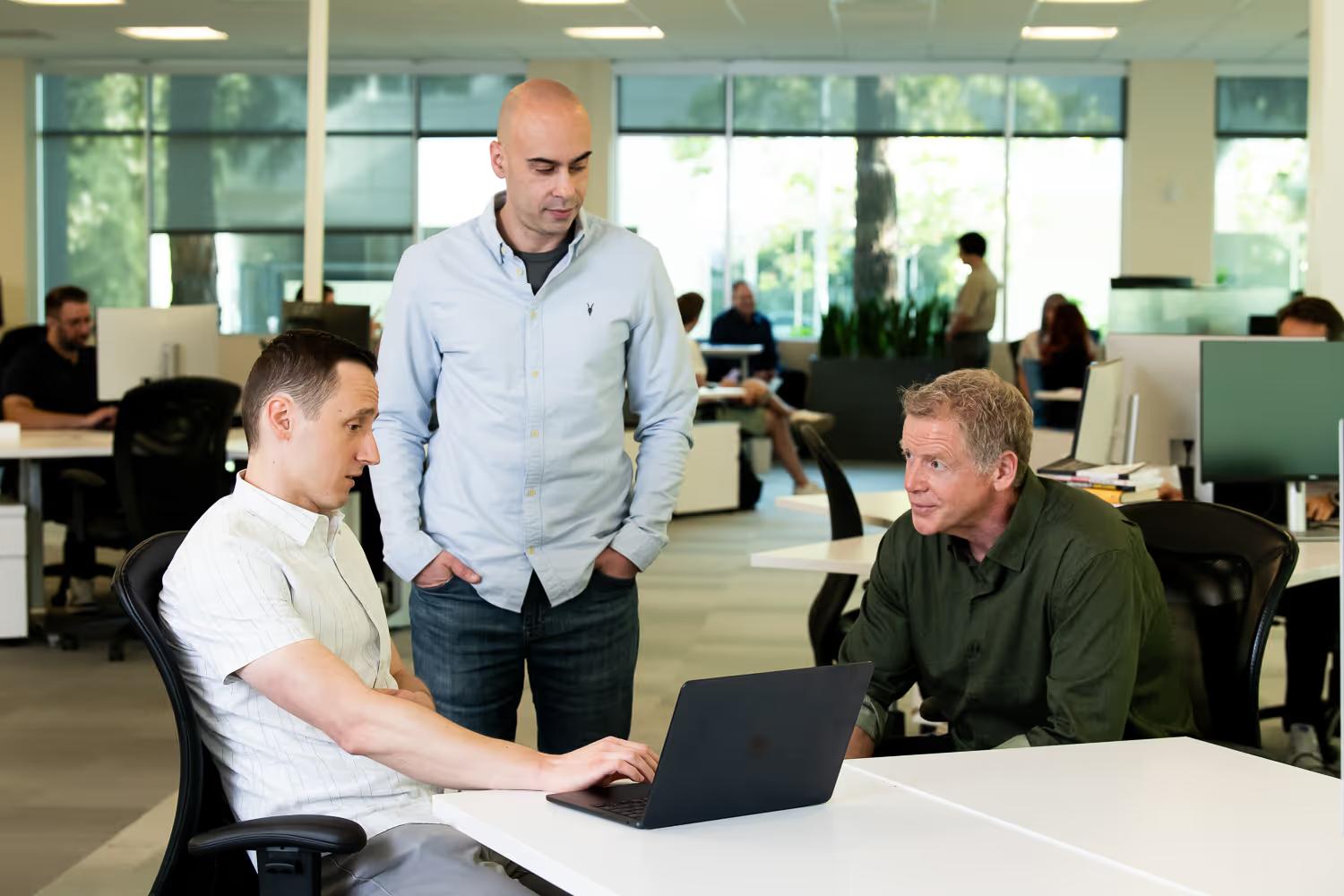 Three men in an office collaborating over a laptop, with one seated typing, one standing, and one seated leaning forward.