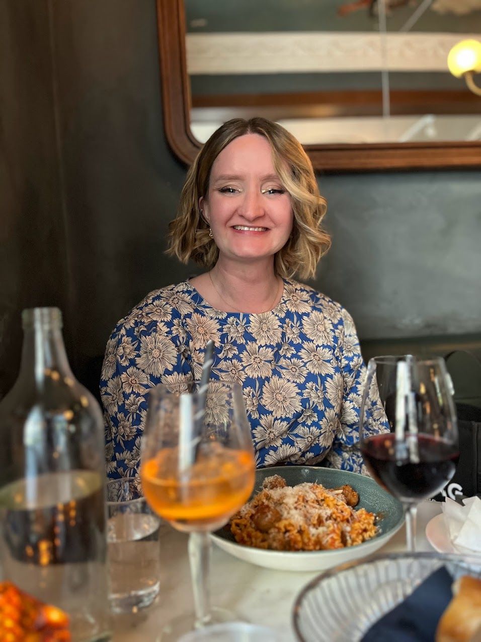 a woman is sitting at a table with a plate of food and two glasses of wine .