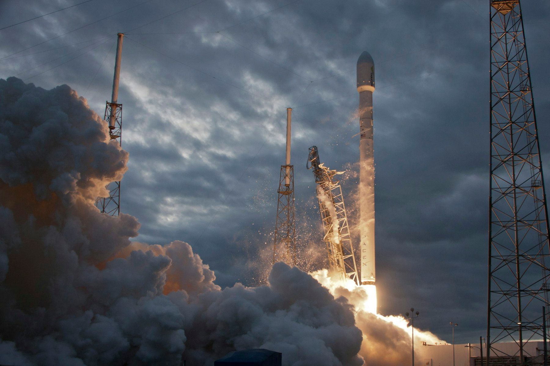 A SpaceX launch vehicle lifting off through smoke at dusk