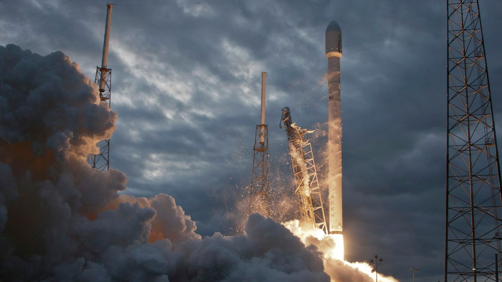 A SpaceX launch vehicle lifting off through smoke at dusk