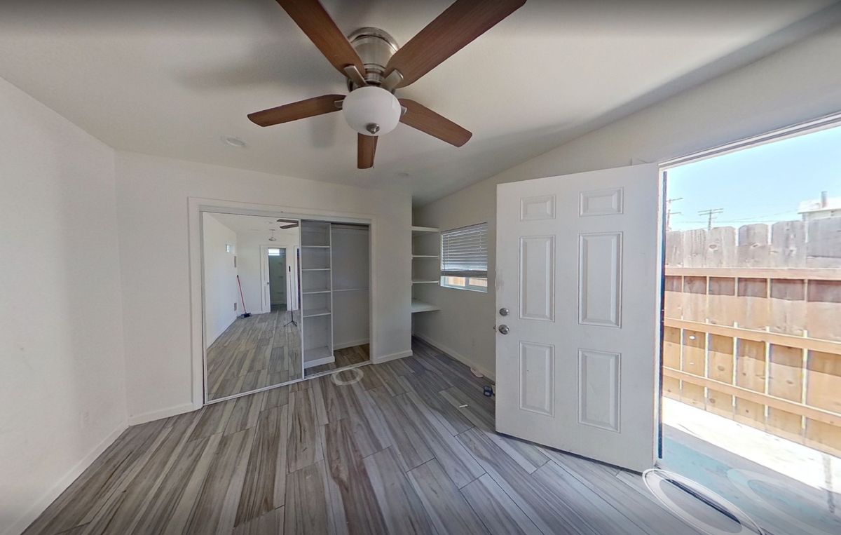 Living area inside an unpermitted garage conversion with a ceiling fan, mirrored closet doors, and an exterior door open to the yard