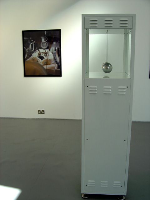 Gallery interior with kinetic sculpture 'One Minute Communication' in foreground with photograph on wall