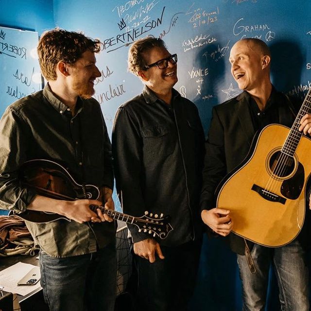 Graham Curry and two bandmates laughing together, holding a fiddle and guitar in front of a graffiti-covered wall.