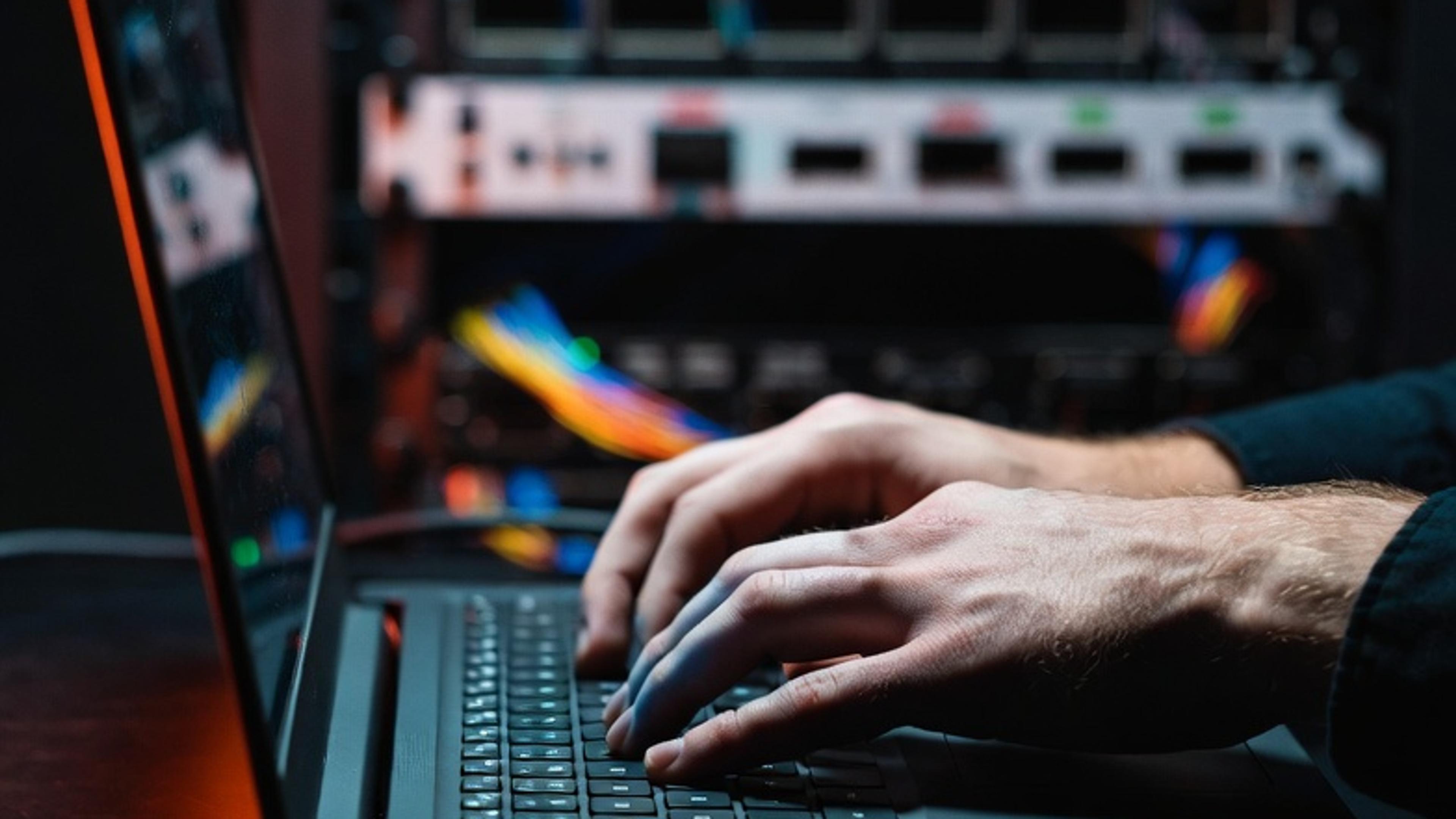 Hands typing on a laptop in front of a server rack with network cables