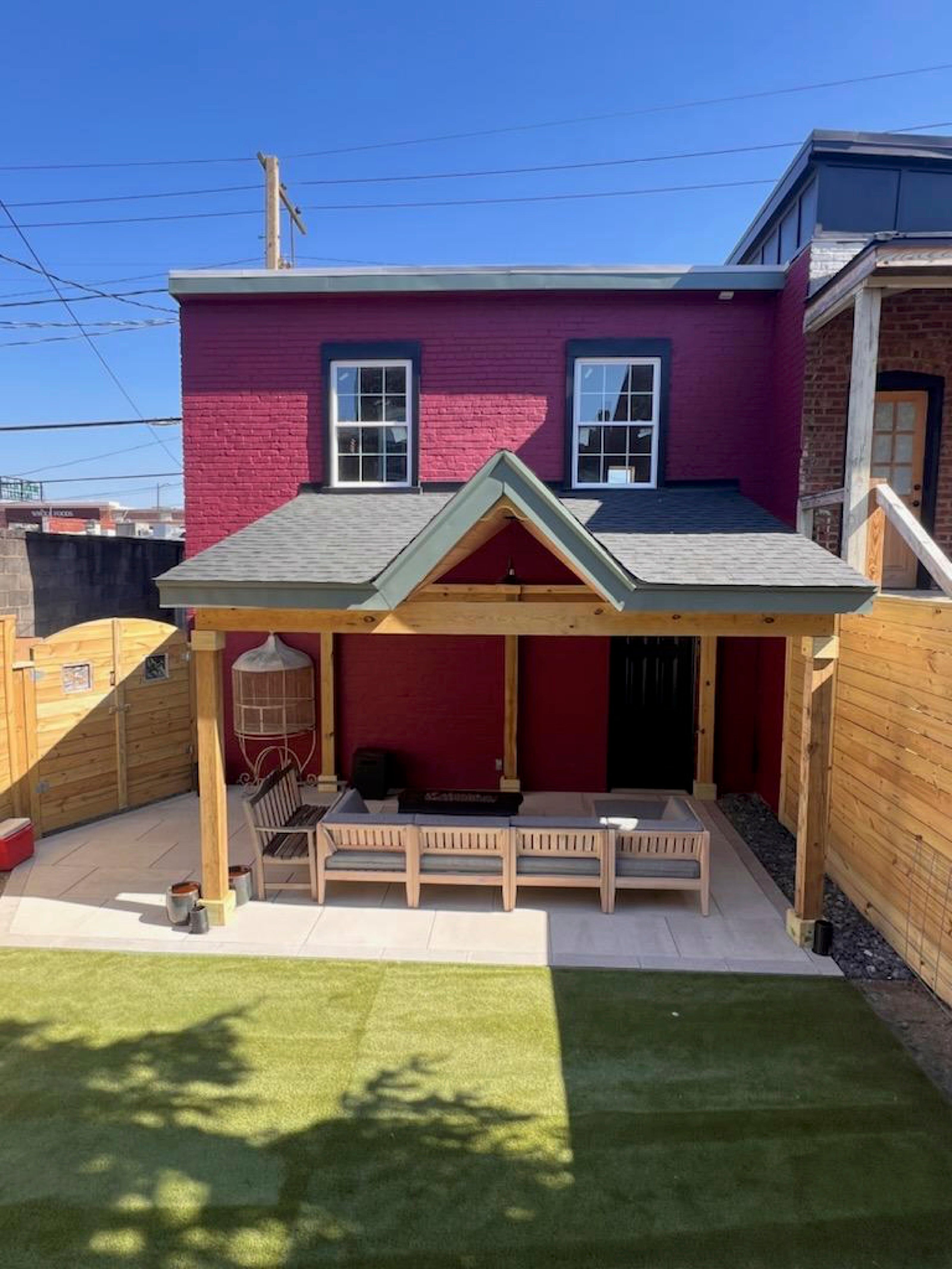 A small brick building with red paint and two windows. A covered patio has wooden seating and a hanging chair. The yard features a wooden privacy fence.