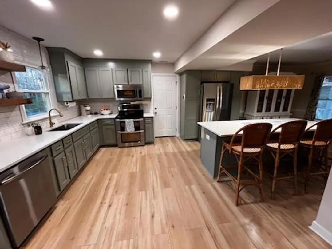 A renovated kitchen with pale green cabinetry surrounding the refridgerator, and an island with a white countertop and bar stools under it. The backsplash is a white subway tile. Wooden shelves stick out of the backsplash on the left side of the sink.