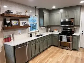 A renovated kitchen with pale green cabinetry and white countertops. The backsplash is a white subway tile. Wooden shelves stick out of the backsplash on the left side of the sink.