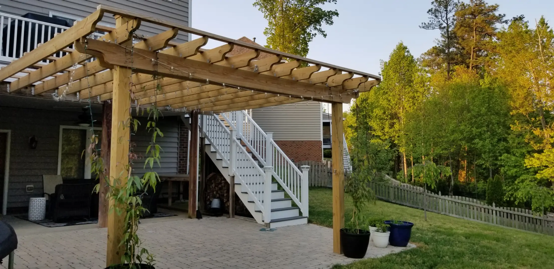 A backyard with a wooden pergola over a paved patio area, featuring a hanging chair and string lights. A white staircase leads to an upper deck.
