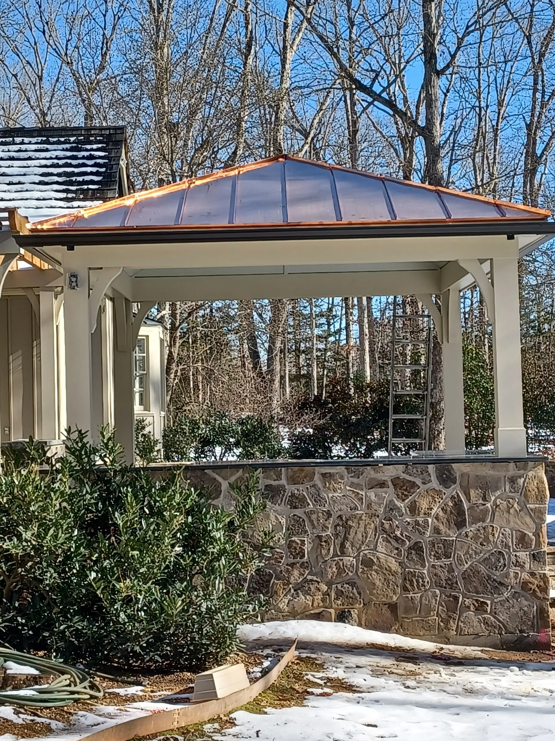 Pavilion with a copper roof and vinyl wrap beams on a stone platform, adjacent to the house, with a path going down around the side of the stone platform