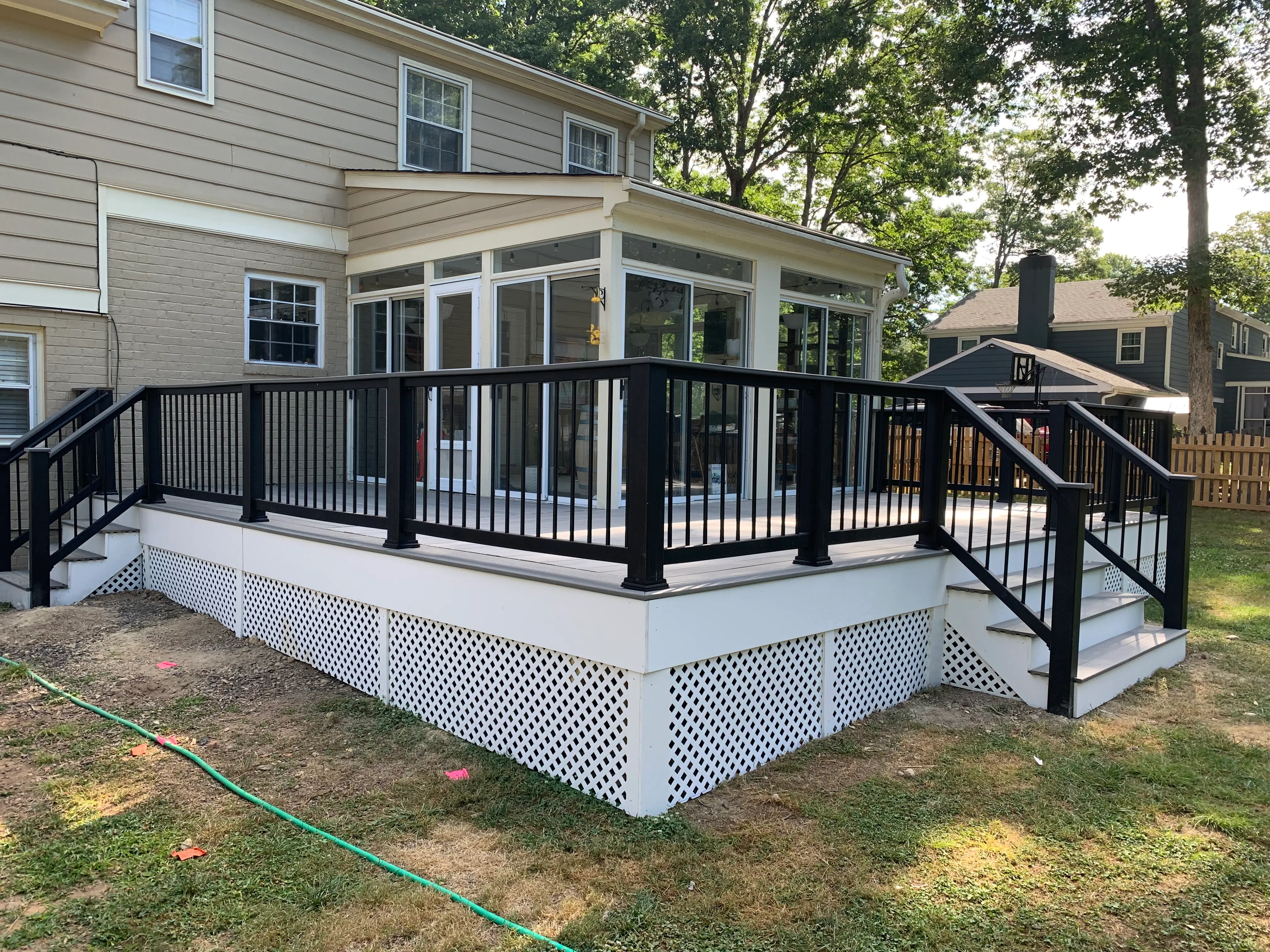 A deck with TimberTech Sea Salt decking and Trex Island Mist Cocktail rail and Trex Enhance composite railings with black aluminum balusters, around the sunroom with two sets of stairs leading the backyard and a white small crossing lattice