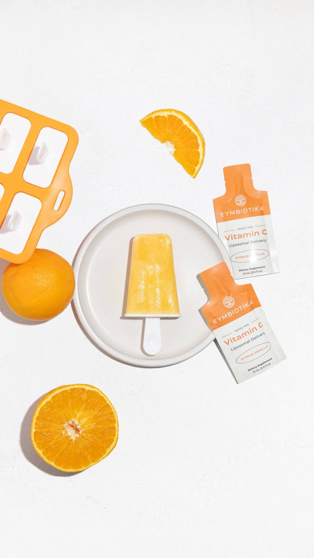 Orange creamsicle on a plate with oranges, a popsicle mold, and Cymbiotika Liposomal Vitamin C packets.