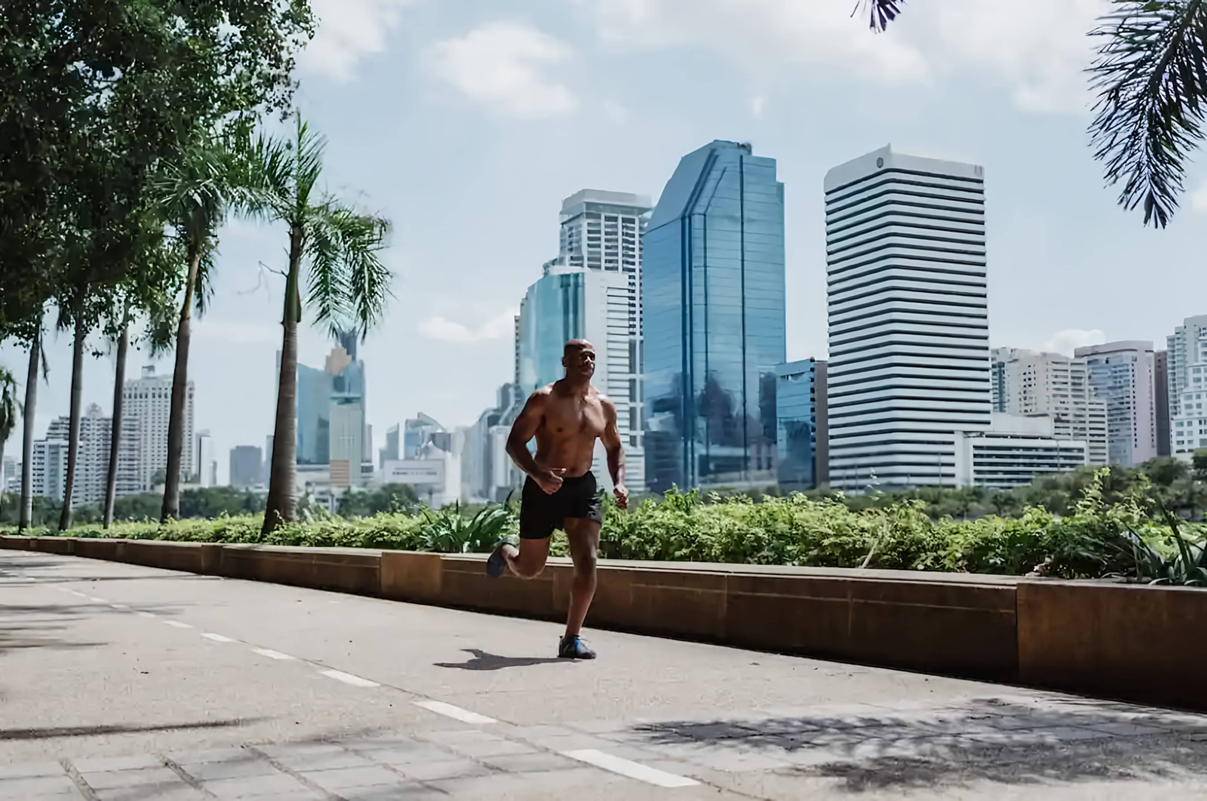 Shirtless man running on a palm-lined city path with high-rise buildings behind him.