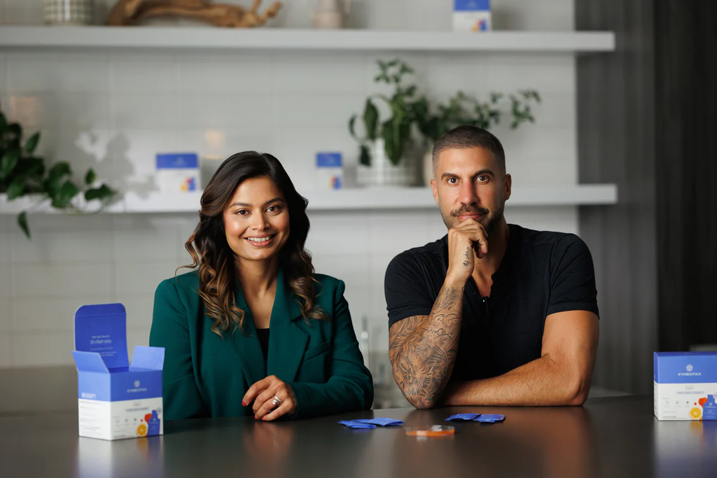 Two people seated behind a table with product packages; shelves and plants are in the background.