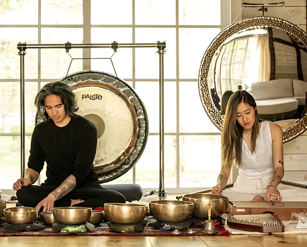 Two people sit among singing bowls, a large gong, and a stringed instrument in a bright room.