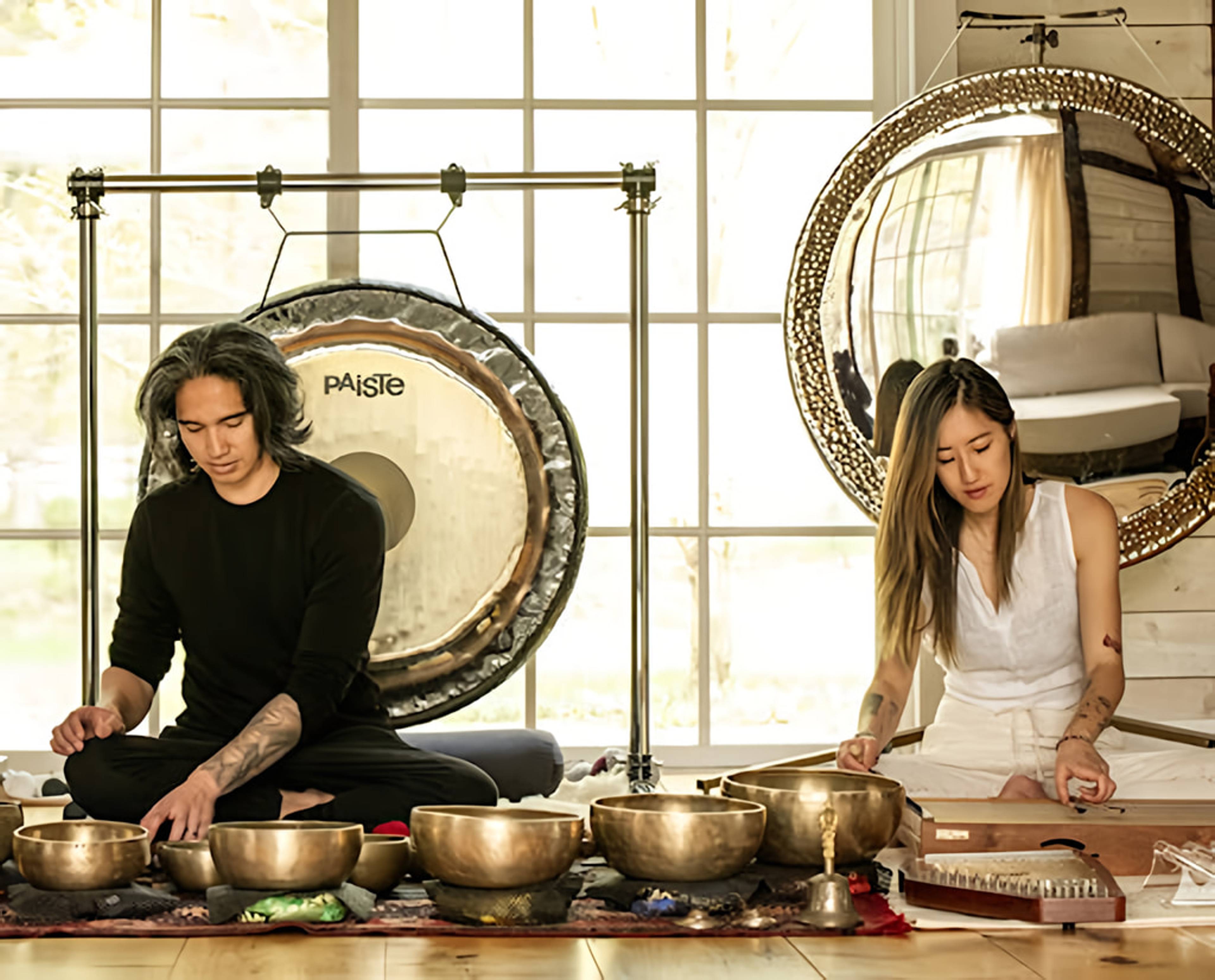 Two people sit among singing bowls, a large gong, and a stringed instrument in a bright room.
